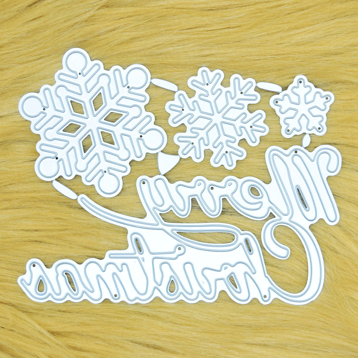 Merry Christmas script and three snowflake metal cutting dies on a tan faux fur background, showing the cutting faces.