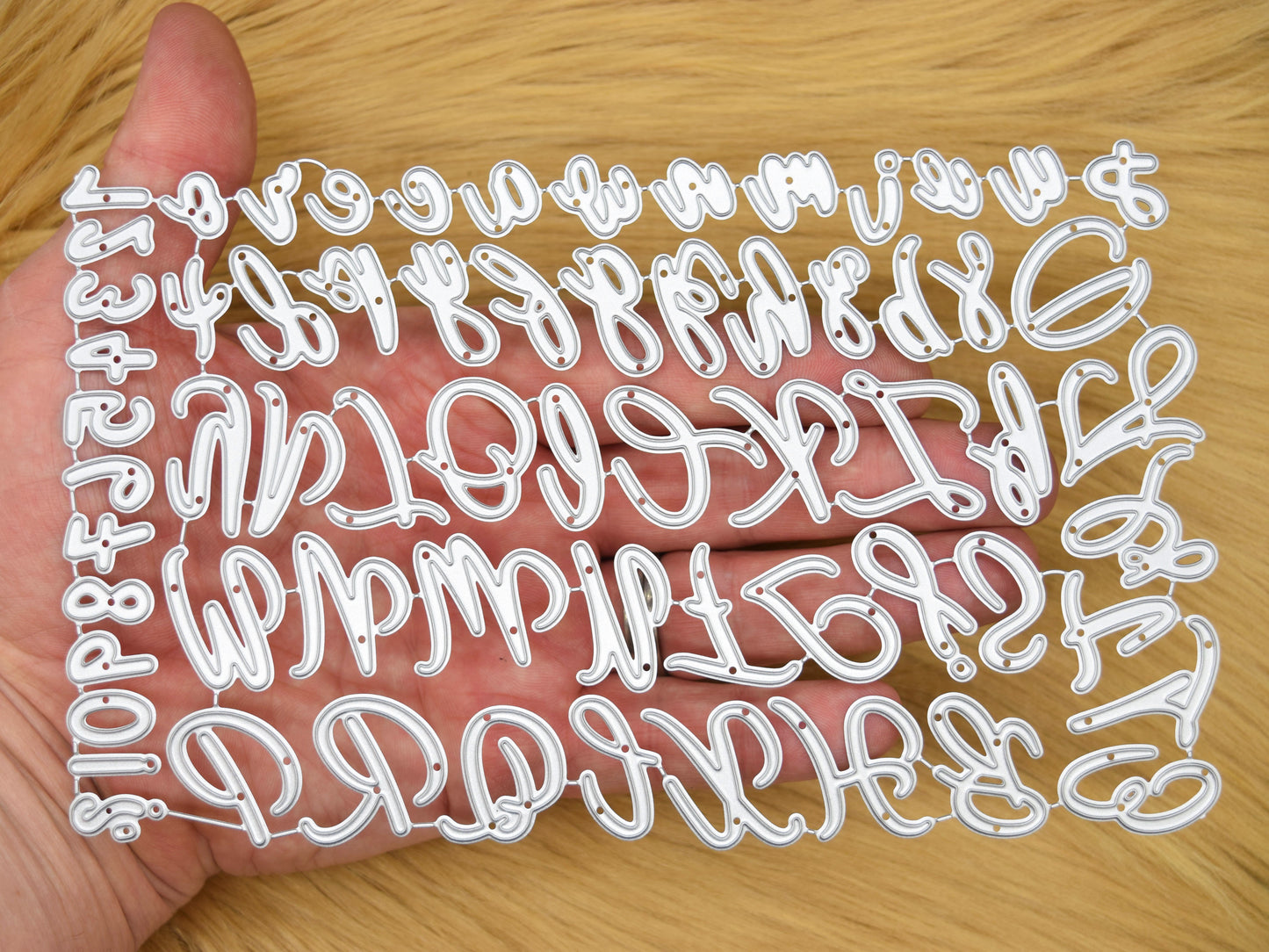 Uppercase & lowercase alphabet and numbers metal cutting dies set shown in hand on a faux fur background to demonstrate the size for card making and papercraft projects.