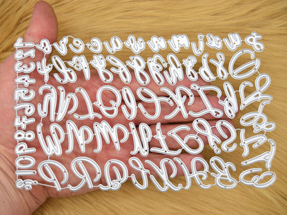 Uppercase & lowercase alphabet and numbers metal cutting dies set shown in hand on a faux fur background to demonstrate the size for card making and papercraft projects.