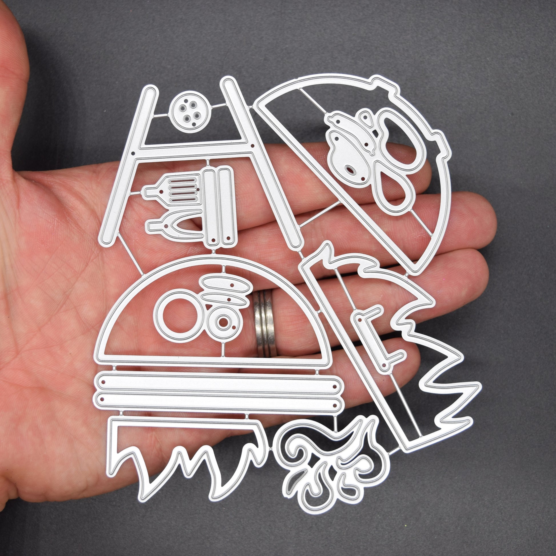 Large BBQ barbecue grill and accessories metal cutting die set shown in hand on dark background for size reference and papercraft use