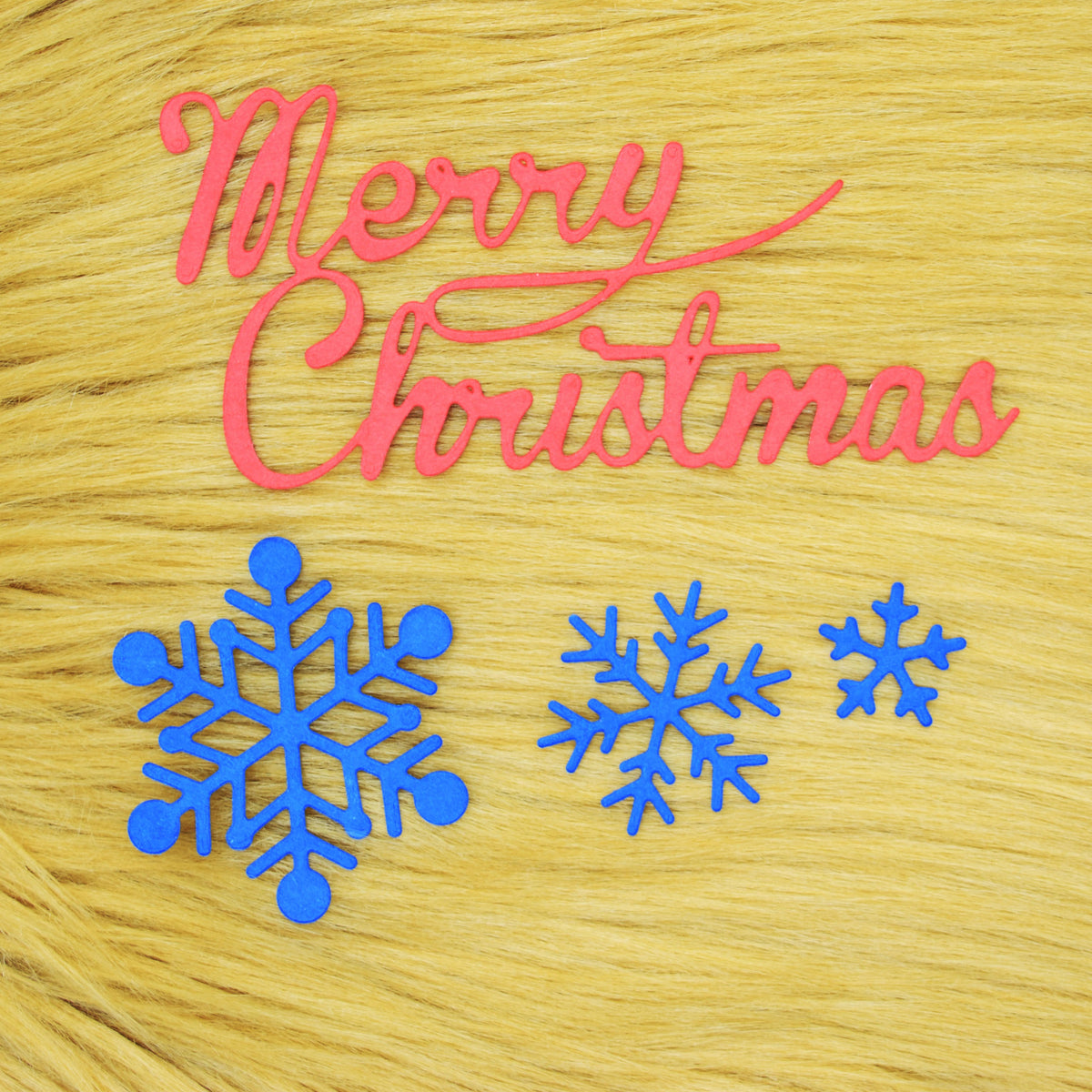 Red Merry Christmas sentiment and three blue snowflake paper cuts arranged on a tan faux fur background for Christmas papercraft.
