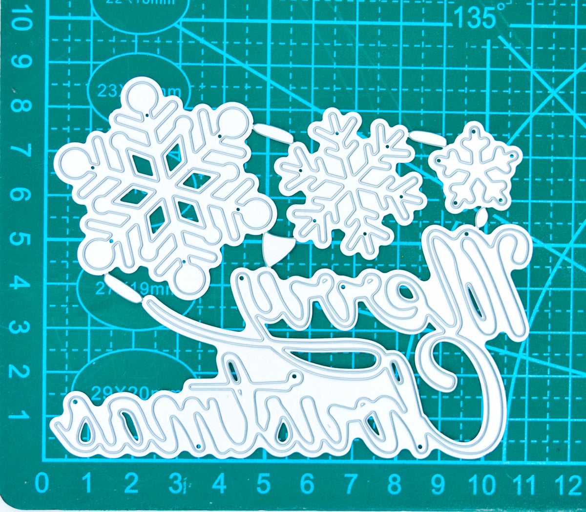 Merry Christmas and snowflake metal cutting dies on a centimetre cutting mat, showing the relative sizes of the four pieces.