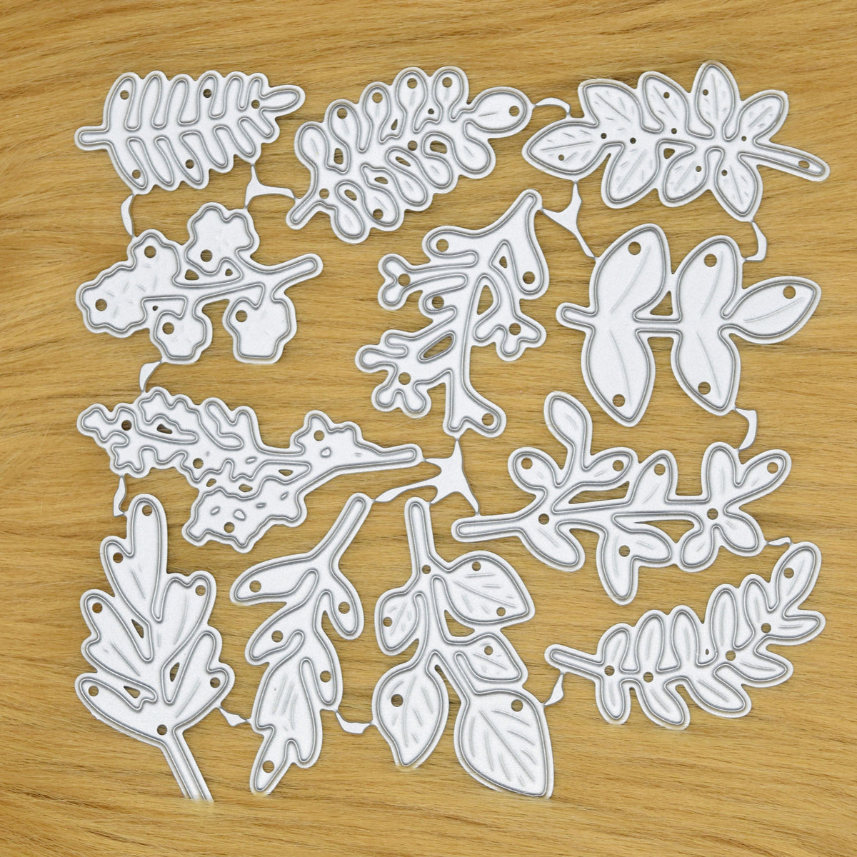 Connected set of 12 leaf and foliage metal cutting dies displayed on a light tan faux fur background