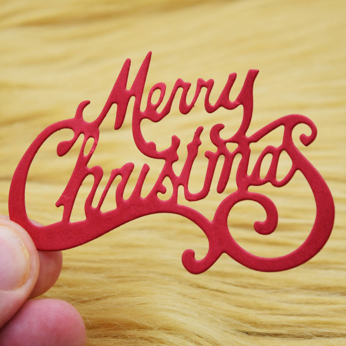 Red Merry Christmas script paper cut held against a tan faux fur background, showing the curled flourishes for festive card making.
