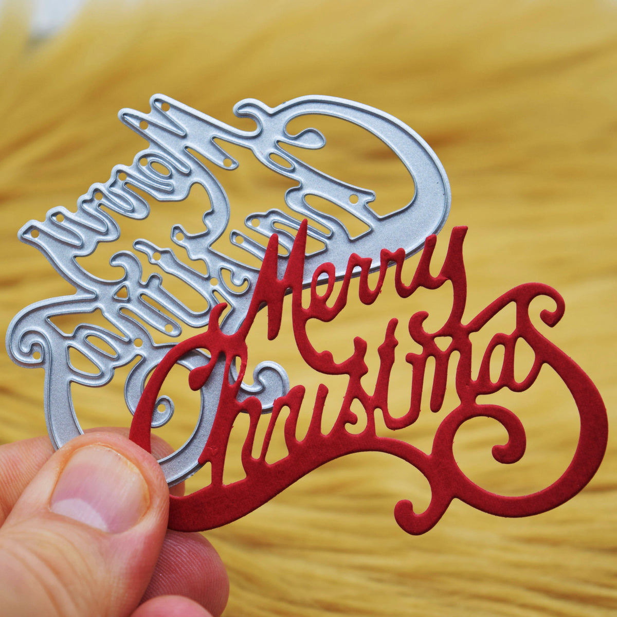 Red Merry Christmas paper sentiment held in front of the metal cutting die against a tan faux fur background for papercraft.