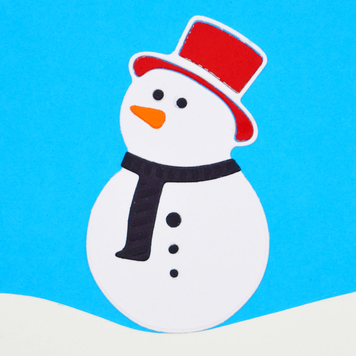 White paper snowman with a red top hat, orange carrot nose and black scarf and buttons, made with metal cutting dies for Christmas card making.