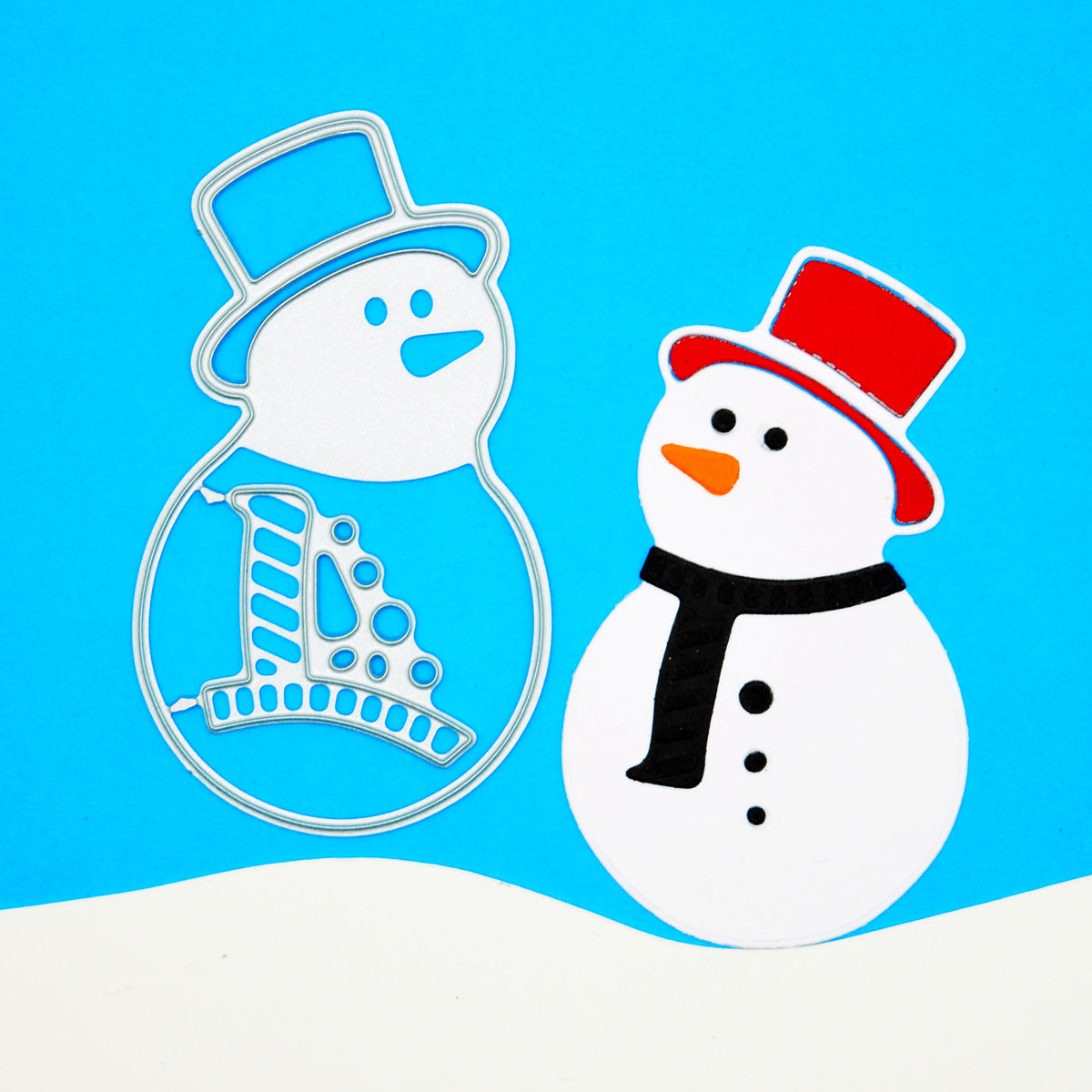Snowman metal cutting dies beside a finished paper snowman on a blue background with a white snowbank for Christmas card making.