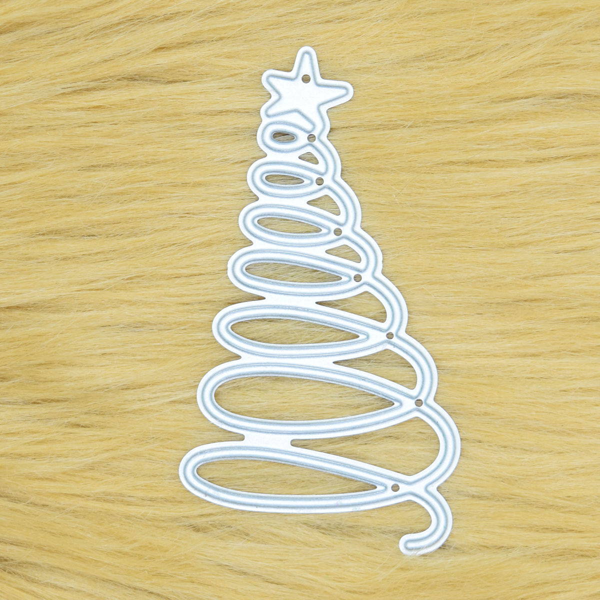 Spiral Christmas tree metal cutting die displayed on a tan faux fur background, showing the looping tree design and star topper.