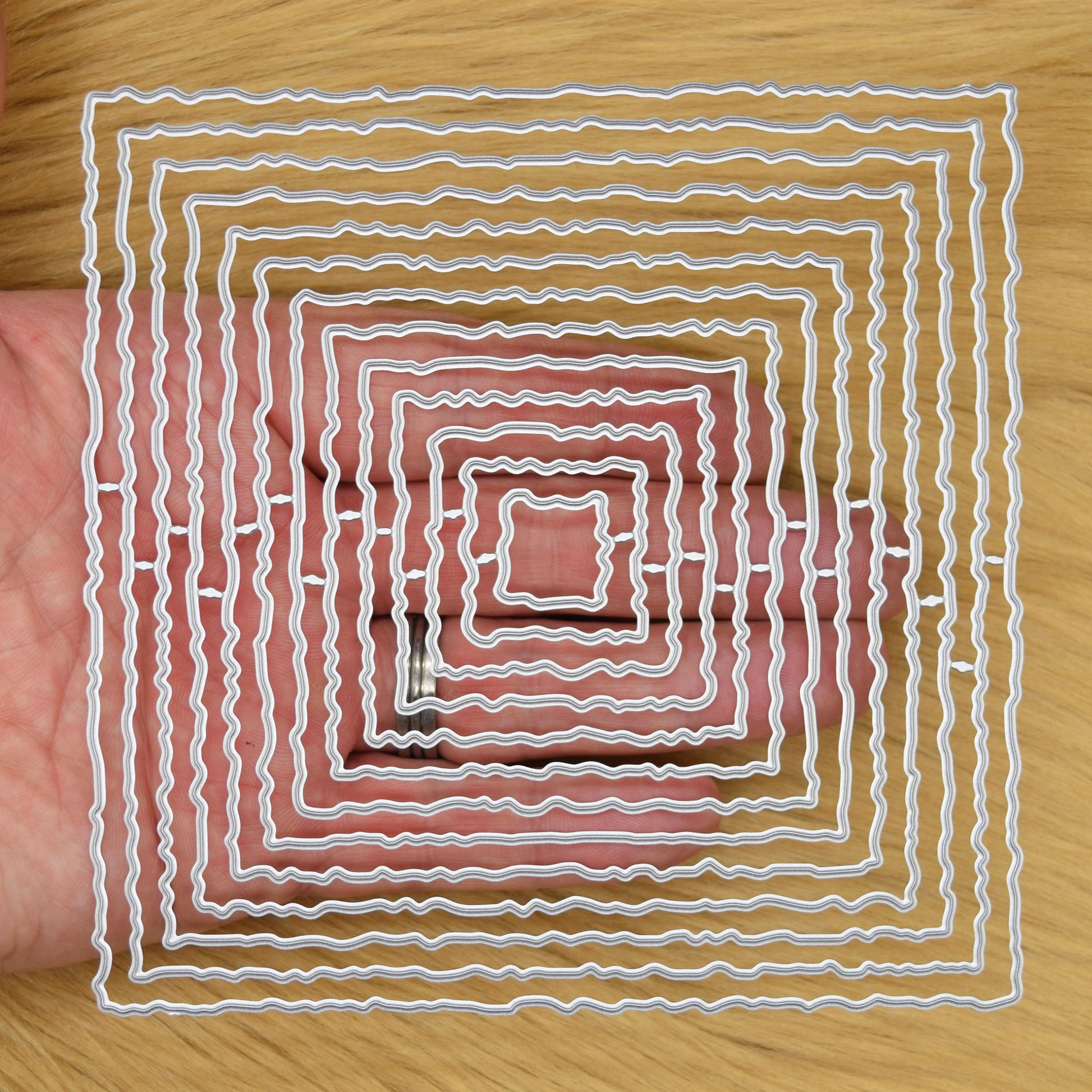 Thirteen graduated torn edge square metal cutting dies held in hand to demonstrate the overall size of the nesting set
